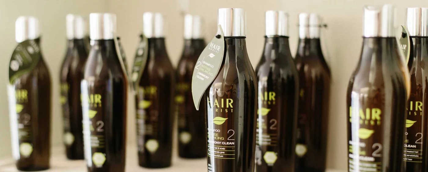 Photo de l’ensemble de la gamme de shampoings bio Hairborist, alignés élégamment sur une étagère en bois naturel, avec des plantes aromatiques et huiles essentielles autour. On distingue clairement les flacons aux étiquettes colorées correspondant à chaque besoin capillaire : Sensity pour les cuirs chevelus sensibles, Daily Clean pour un usage fréquent, Harmony Clean pour réguler le sébum, Color Clean pour cheveux colorés, Men Tonic pour les hommes actifs, et Naturalys pour équilibrer le microbiote. L’image évoque l’univers naturel, écoresponsable et artisanal de la marque Hairborist, en harmonie avec les valeurs de La Maison du Coloriste.