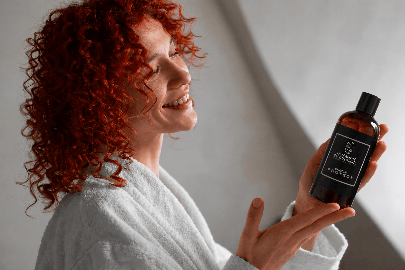 Femme aux cheveux bouclés roux tenant le shampoing Chroma Protect La Maison du Coloriste, soin cheveux colorés